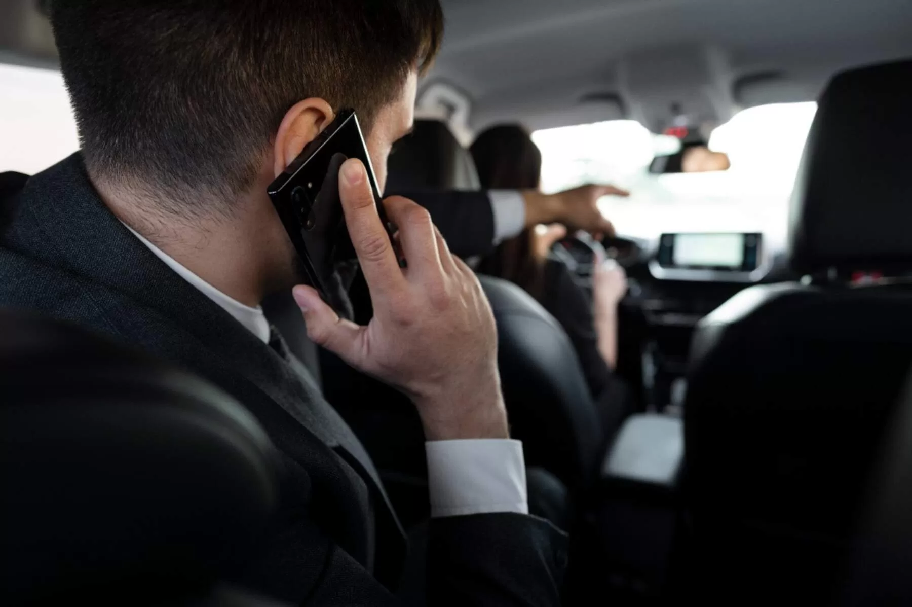 Passenger in a suit talking on the phone inside a taxi, while the driver is driving.